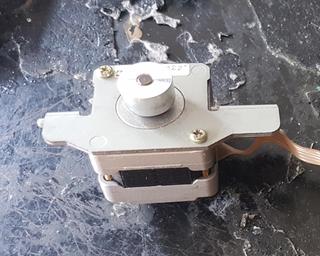 Small stepper motor on a stamped metal bracket with a flat flexible cable coming out of it.